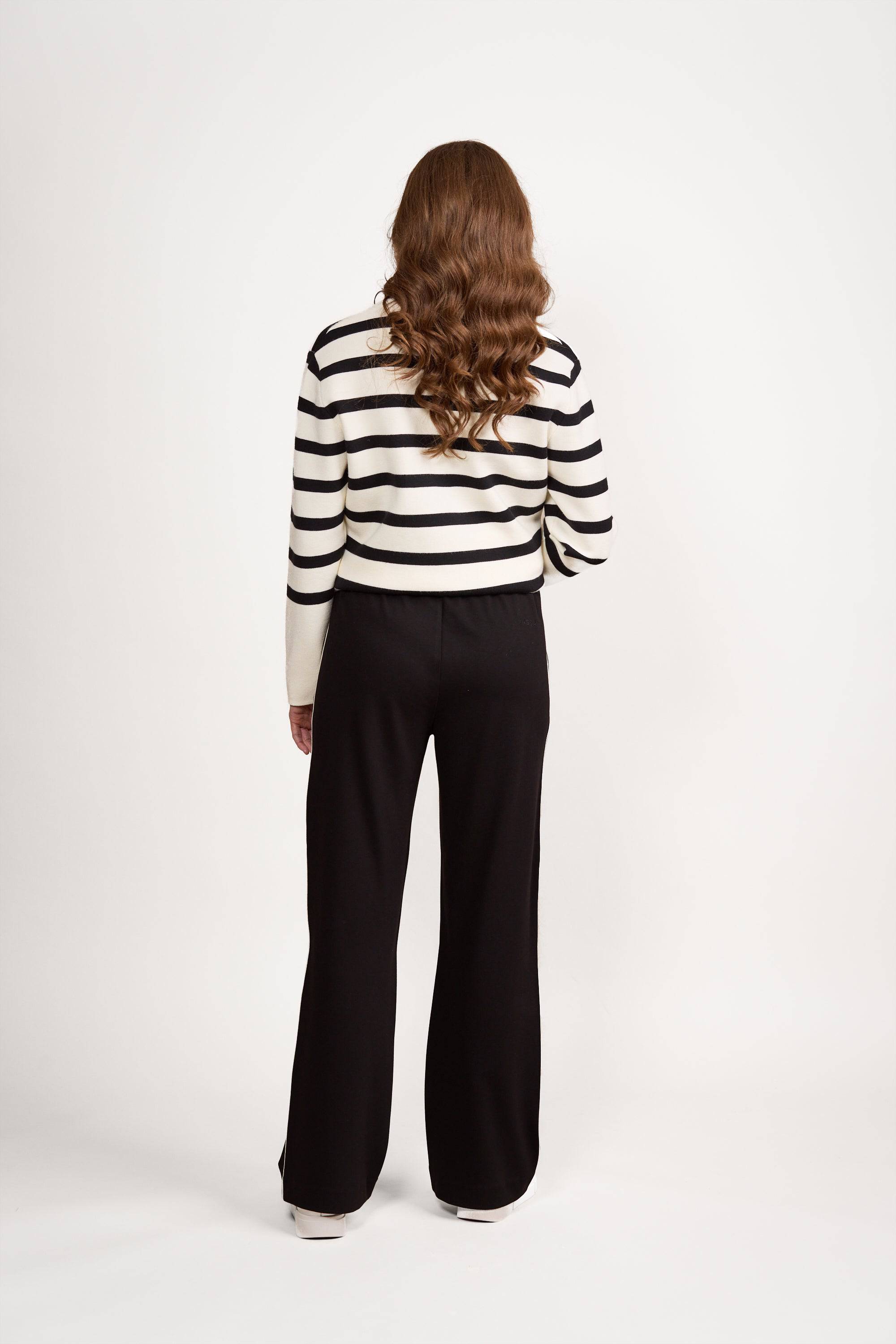 A person with long, wavy brown hair, facing away, wears a white sweater with black stripes and Vassalli’s Weekend Pant - Black 2.0, loose wide-leg stretch pants, against a plain white background.