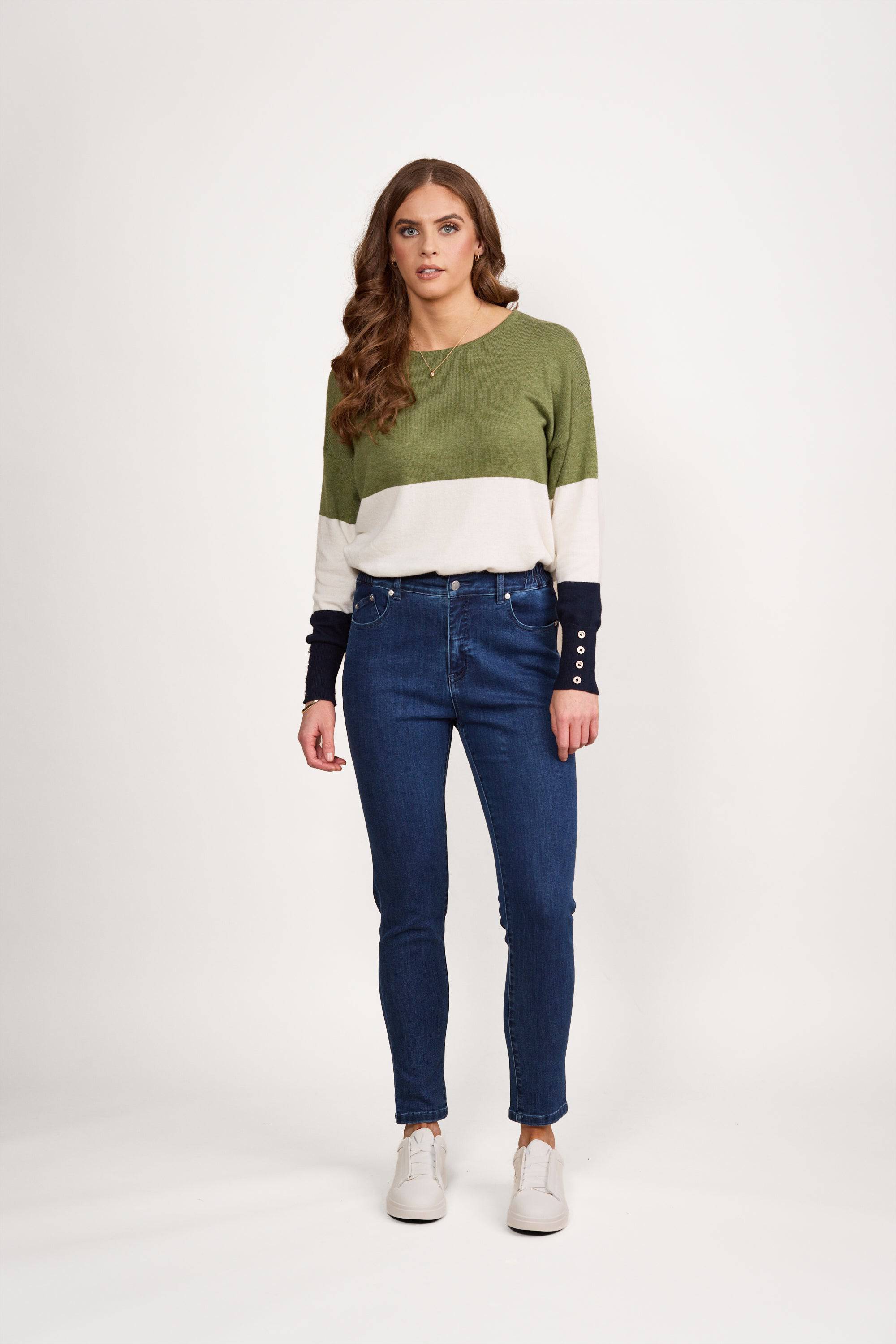 A woman with long wavy brown hair stands facing forward against a white background, wearing a green, white, and navy color-block sweater, Vassalli Mode Jean - Blue Denim, and white sneakers. She has a neutral expression.