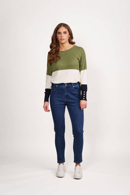 A woman with long wavy brown hair stands facing forward against a white background, wearing a green, white, and navy color-block sweater, Vassalli Mode Jean - Blue Denim, and white sneakers. She has a neutral expression.