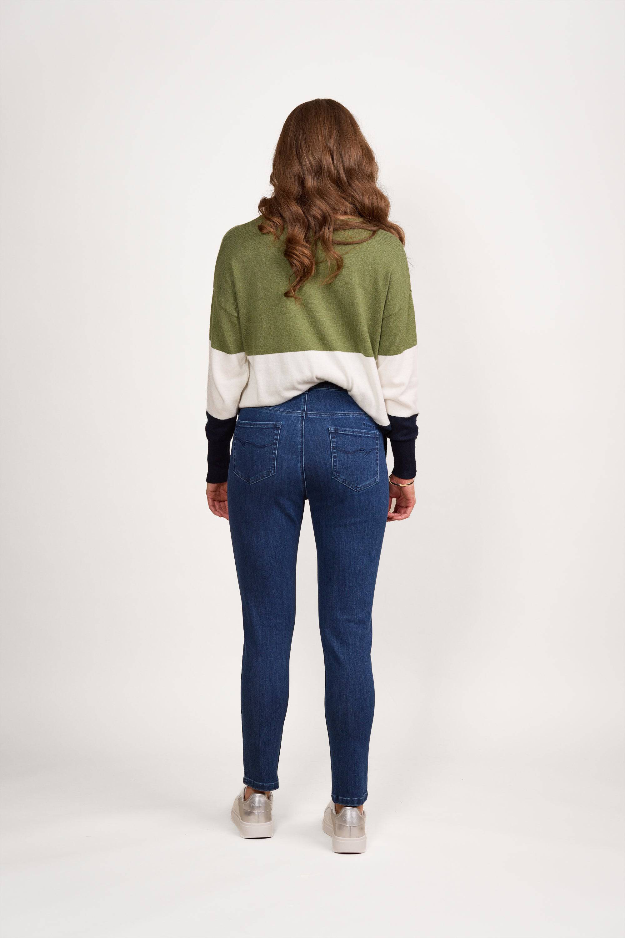 A person with long, wavy brown hair stands facing away, wearing a green and white color-block sweater, Vassalli Mode Jean in blue denim, and silver sneakers against a plain white background.