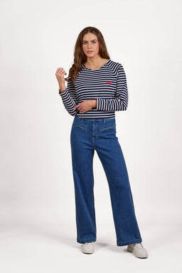 A woman with long brown hair stands against a white background, wearing a navy and white striped long-sleeve top, Vassalli Peace Jean - Blue Wash high-waisted wide leg jeans, and white shoes. She has a neutral expression.