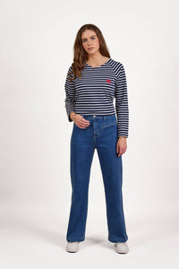 A woman with long brown hair stands facing forward, wearing a navy and white striped long-sleeve shirt, Vassalli Peace Jean - Blue Wash wide leg jeans in stretch fabric, and white shoes, set against a plain white background.
