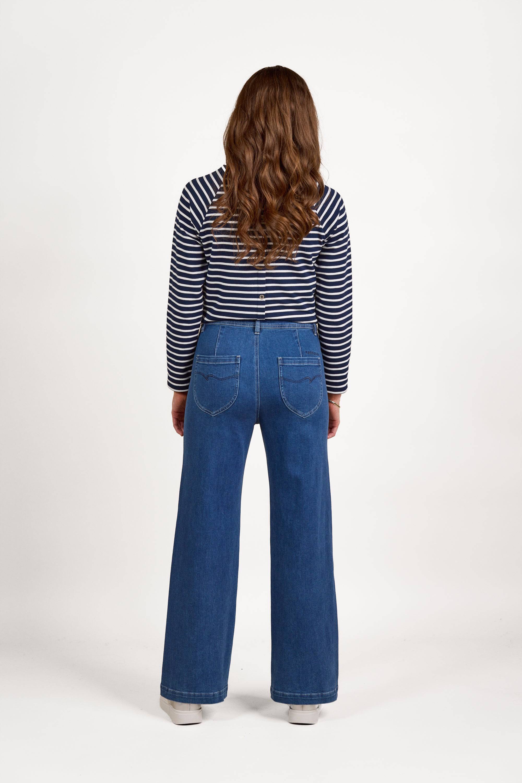 A person with long, wavy brown hair faces away, wearing a navy and white striped long-sleeve shirt and Vassalli Peace Jean in Blue Wash, set against a plain white background.