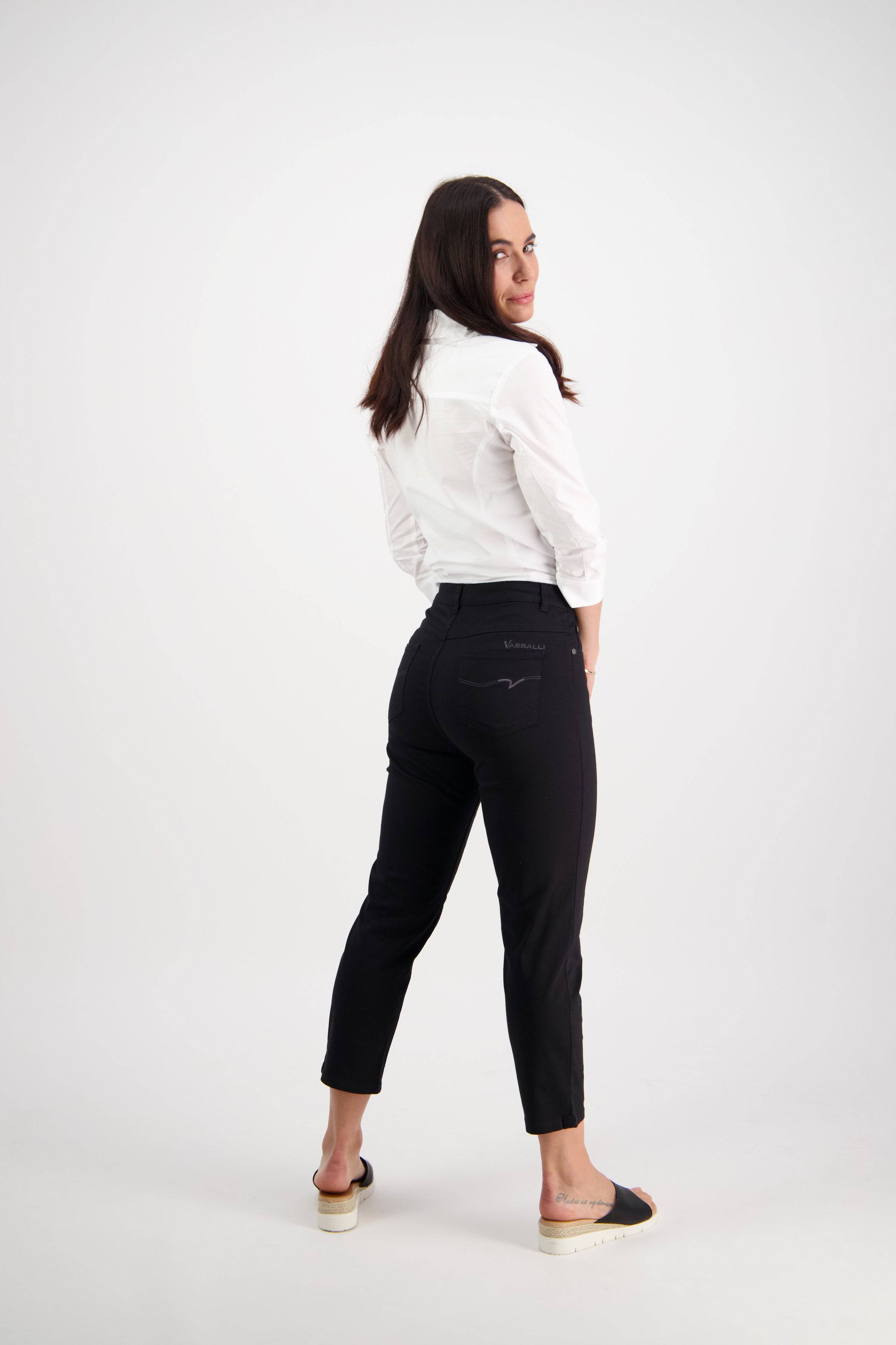 A woman with long dark hair models the Vassalli Sara 7/8 Pant in black, paired with a white shirt. She stands turned to the side, looking over her shoulder against a plain white background.