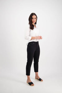 A woman with long dark hair stands against a white background, wearing the Vassalli Sara 7/8 Pant in Black paired with a white button-up shirt and black open-toe sandals, smiling slightly with her hands clasped in front.