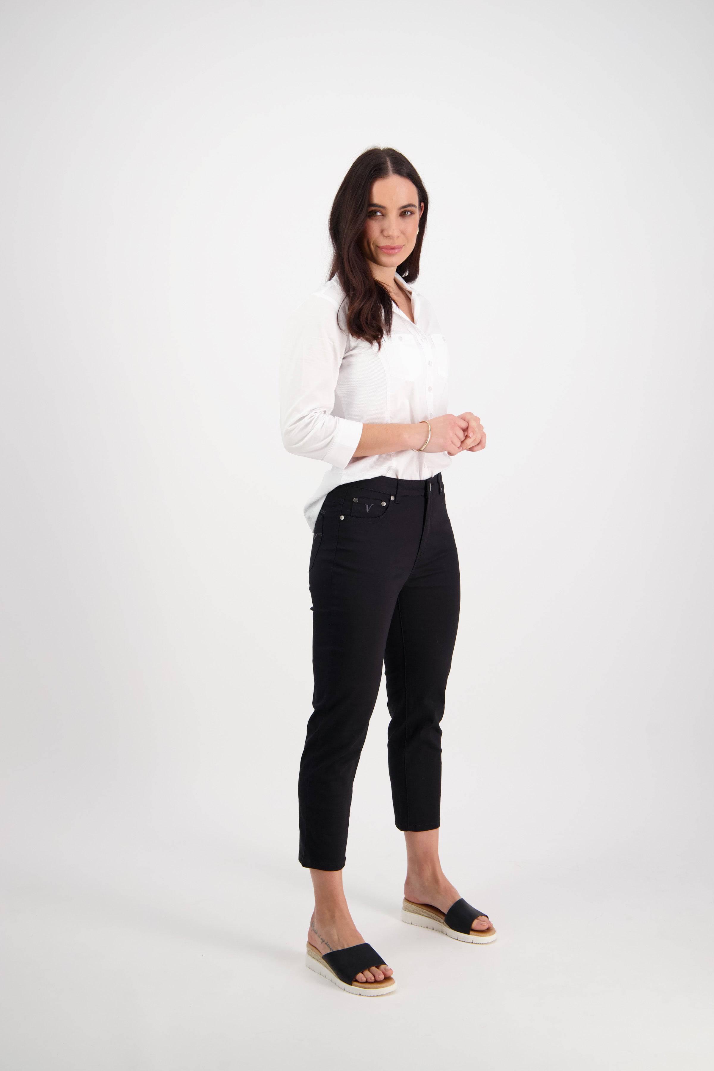 A woman with long dark hair stands against a white background, wearing the Vassalli Sara 7/8 Pant in Black paired with a white button-up shirt and black open-toe sandals, smiling slightly with her hands clasped in front.