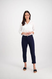 A woman with long dark hair, smiling and clasping her hands, wears a white shirt and Vassalli's Sara 7/8 Pant in navy skinny fit, styled with black open-toe sandals against a plain white background.