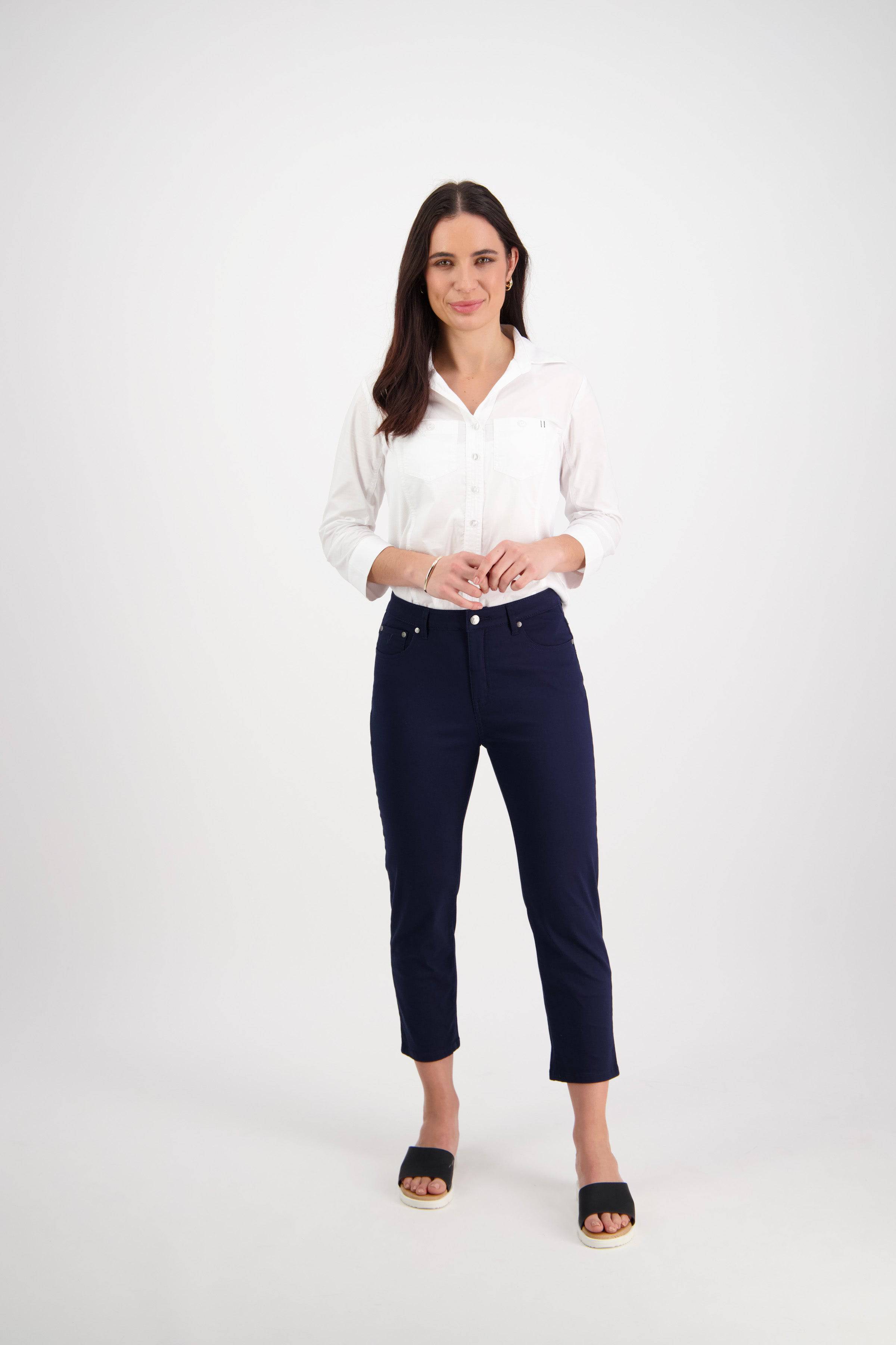 A woman with long dark hair, smiling and clasping her hands, wears a white shirt and Vassalli's Sara 7/8 Pant in navy skinny fit, styled with black open-toe sandals against a plain white background.