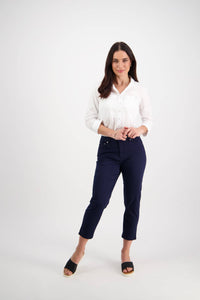 A woman with long dark hair stands against a white background, smiling softly with her hands clasped. She wears a white button-up shirt, Vassalli Sara 7/8 Pant in navy, and black slide sandals.