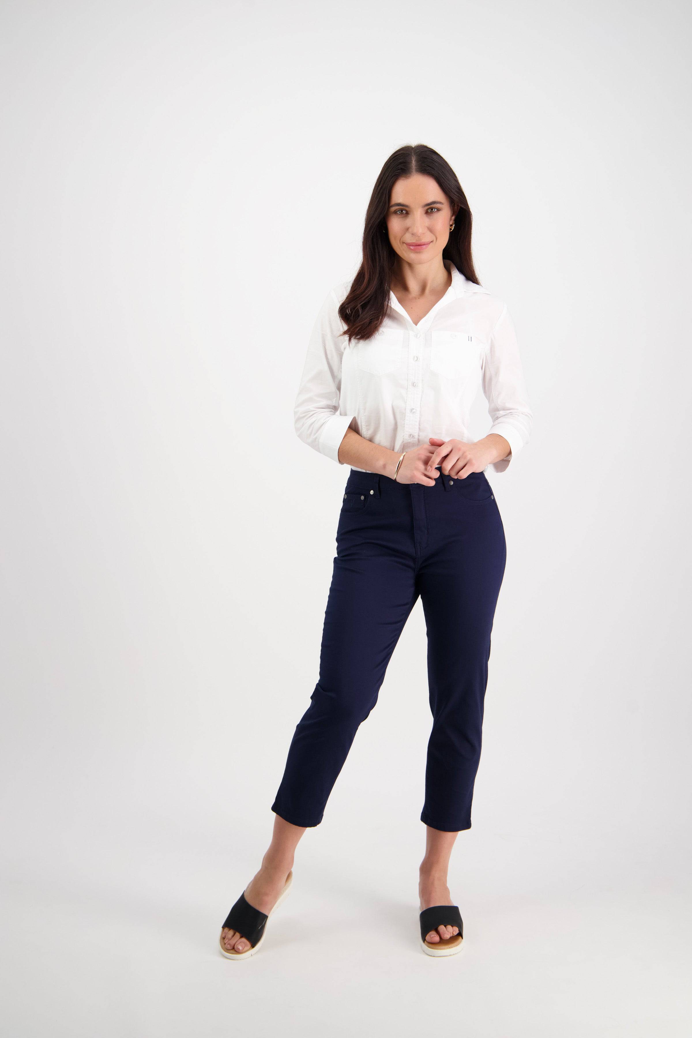 A woman with long dark hair stands against a white background, smiling softly with her hands clasped. She wears a white button-up shirt, Vassalli Sara 7/8 Pant in navy, and black slide sandals.