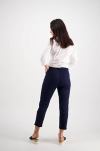 A woman with long dark hair, in a white blouse and Vassalli Sara 7/8 Pant - Navy, paired with black slip-on shoes, stands with her back to the camera against a plain white background.