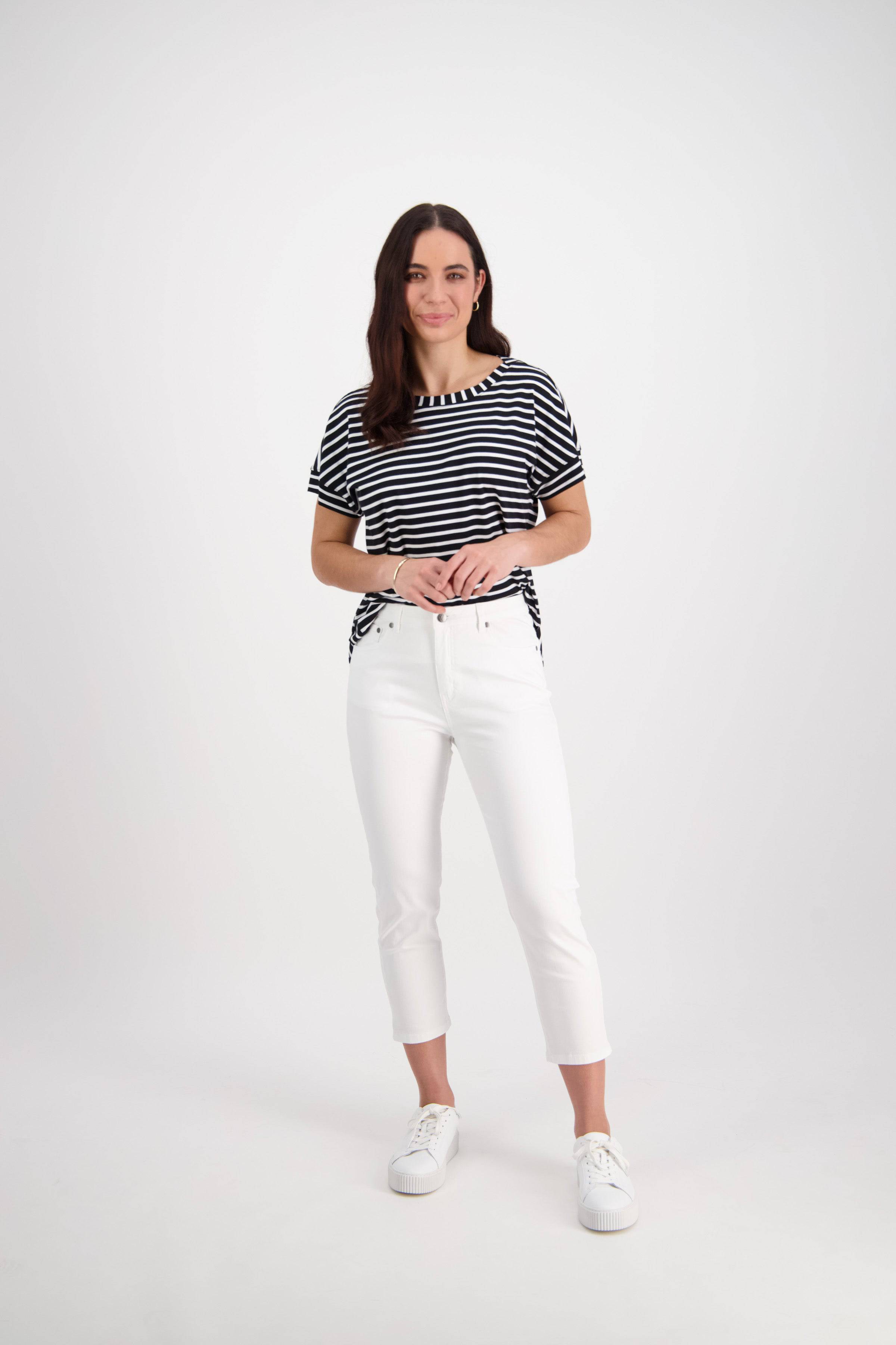 A woman with long dark hair stands against a plain white background, smiling slightly with her hands clasped, wearing the Vassalli Sara 7/8 Pant in White, a black and white striped t-shirt, and white sneakers.