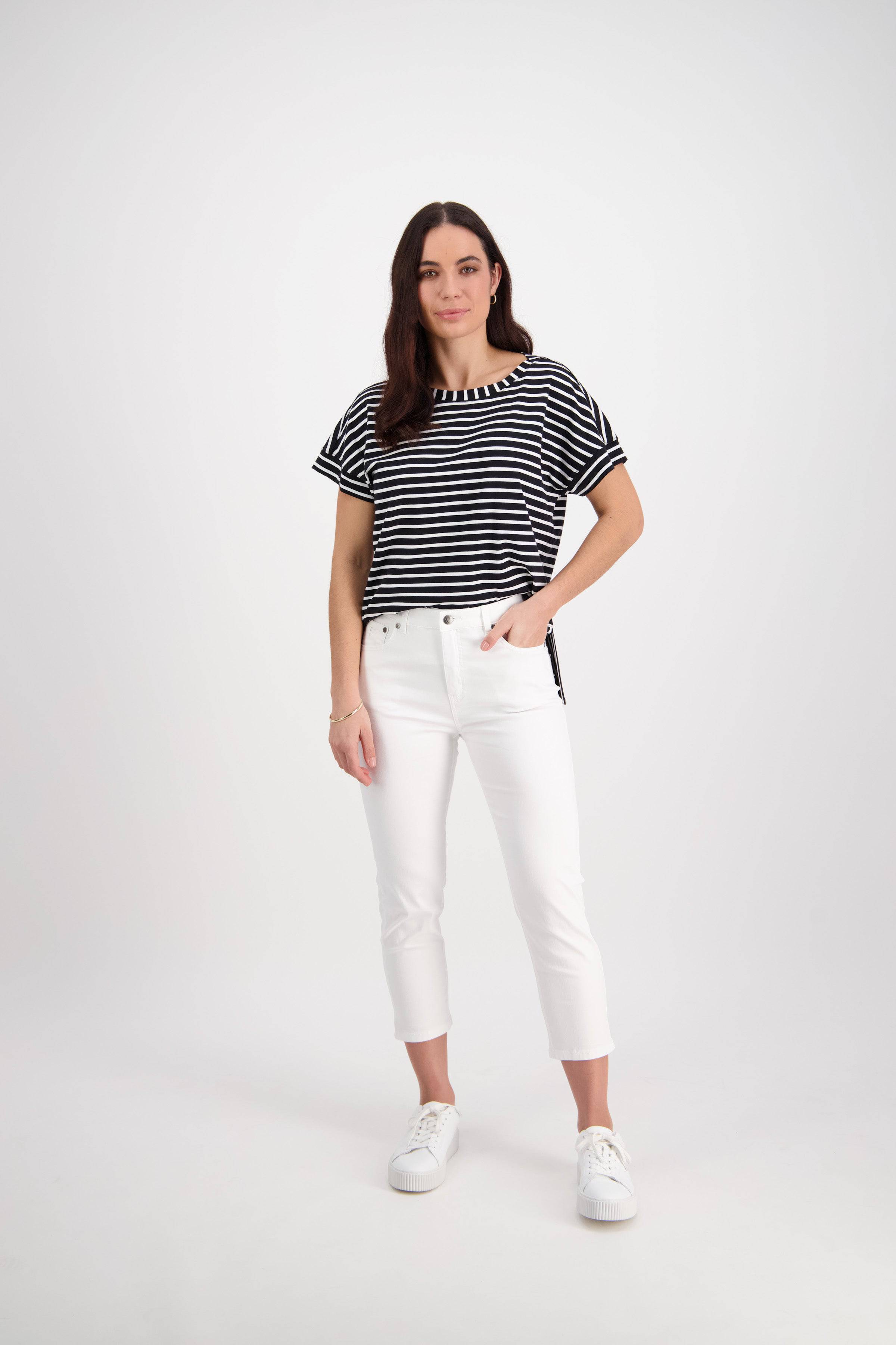 A woman with long brown hair and a neutral expression stands against a white background, wearing Vassalli's Sara 7/8 Pant in white, a black-and-white striped short-sleeve top, and white sneakers, with one hand in her pocket.