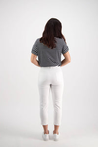 A woman with long brown hair faces away, wearing a black and white striped t-shirt, Vassalli's Sara 7/8 Pant in White, and white sneakers against a plain white background.