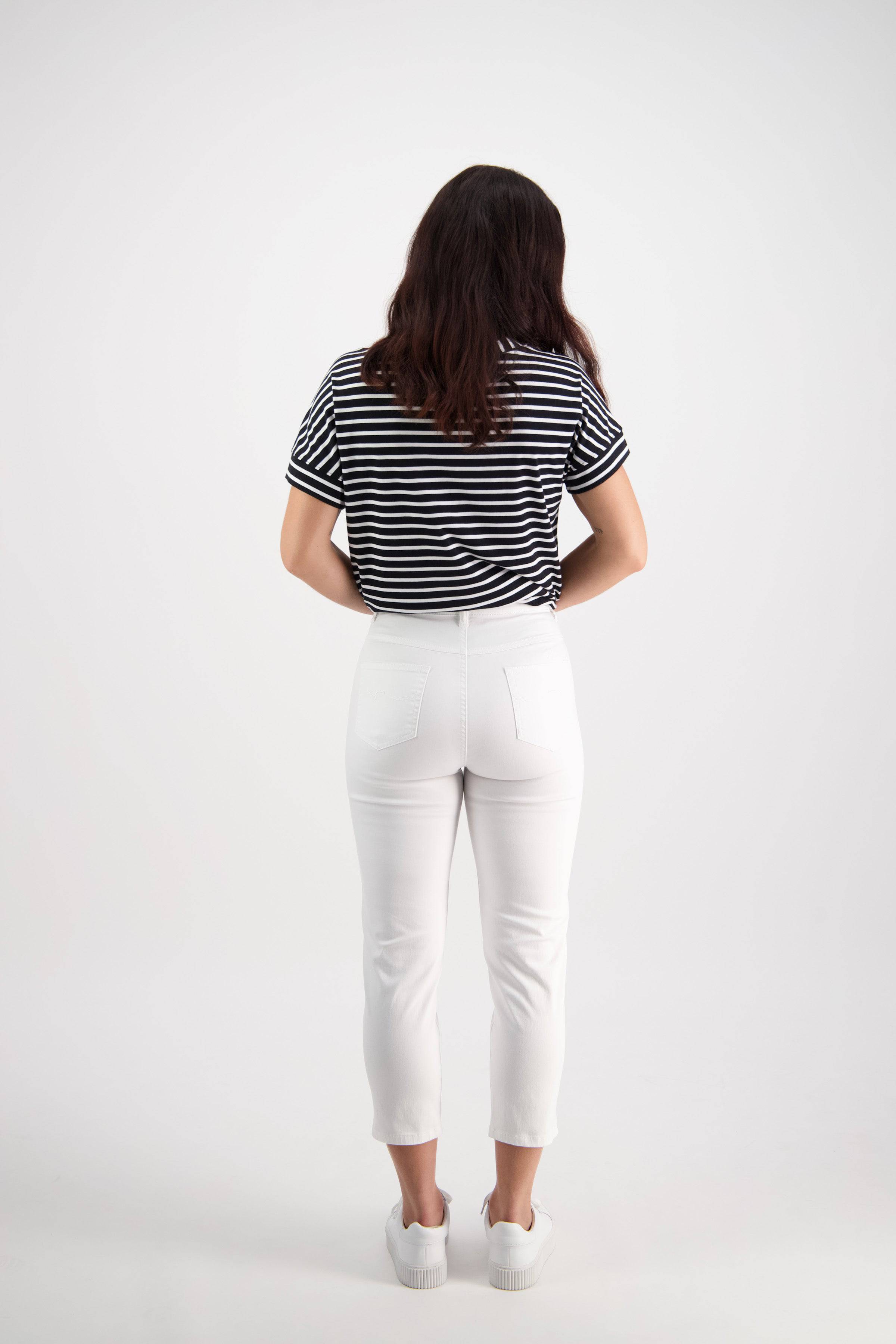 A woman with long brown hair faces away, wearing a black and white striped t-shirt, Vassalli's Sara 7/8 Pant in White, and white sneakers against a plain white background.