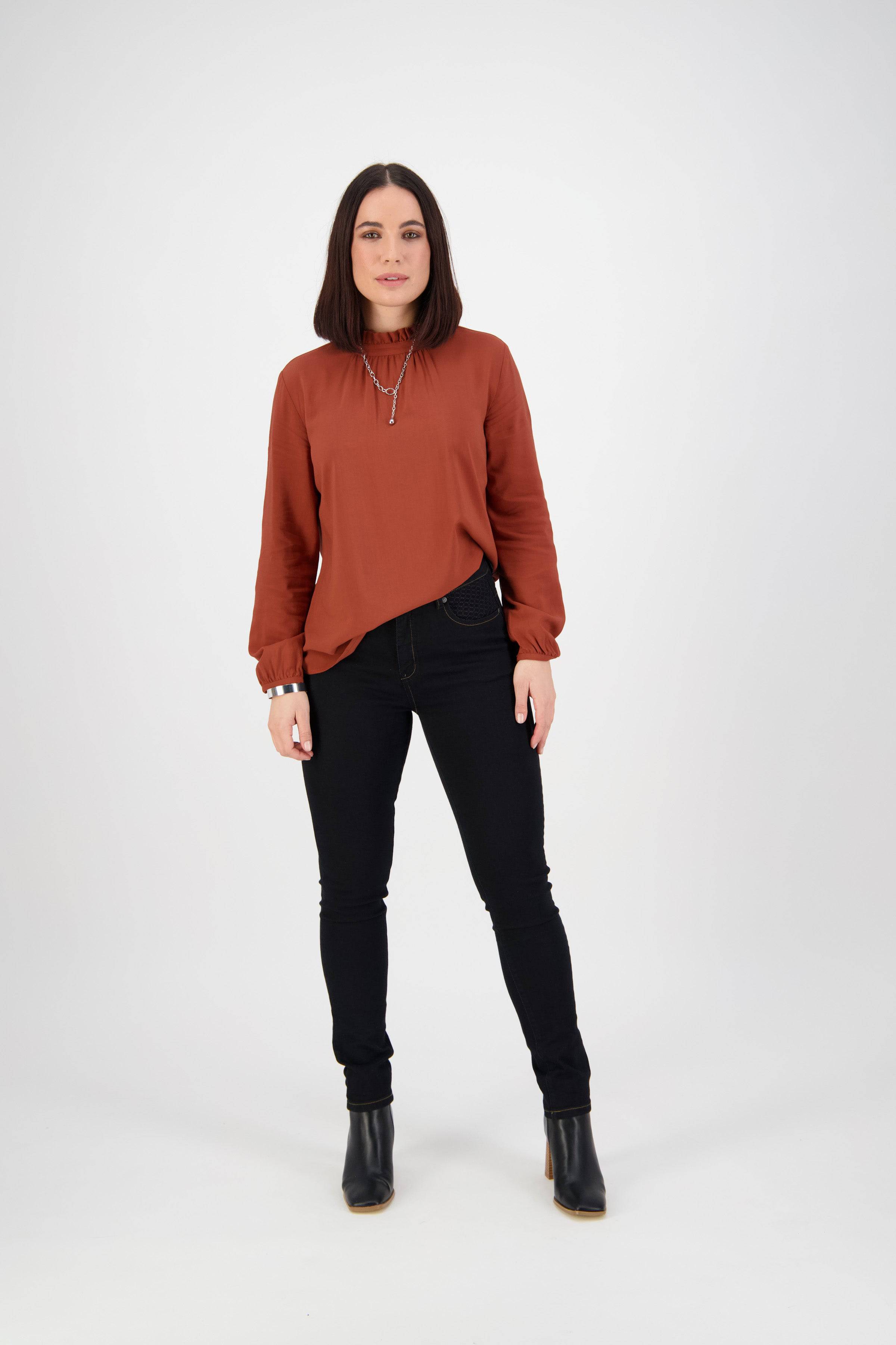 A woman with straight dark hair wears a rust blouse, the Vassalli Reign Jean in Black Denim, and black ankle boots. She stands facing forward against a plain white background with a neutral expression.