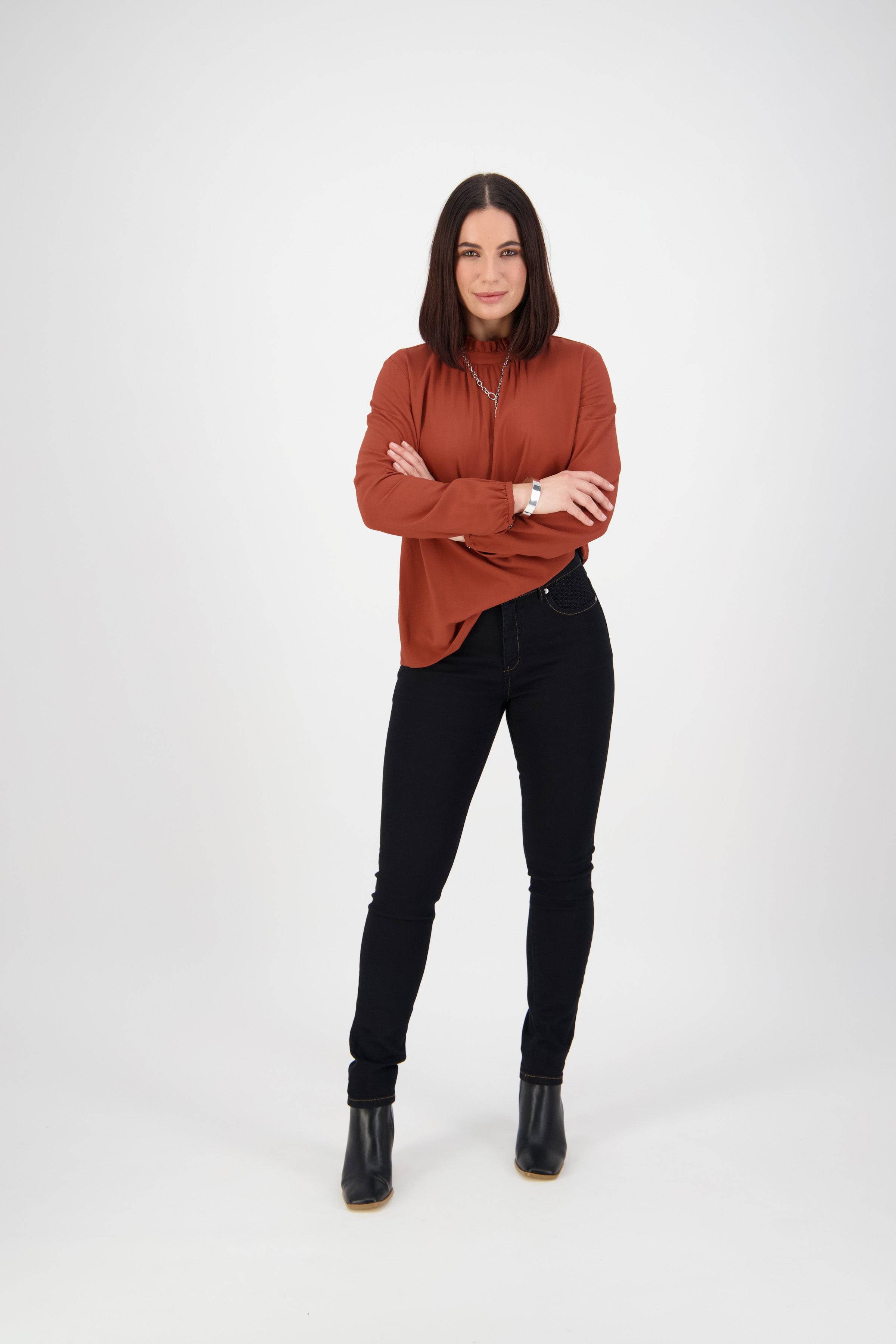 A woman with straight dark hair stands confidently with arms crossed, wearing a rust-colored blouse, Vassalli Reign Jean in black denim, and black ankle boots against a plain white background.