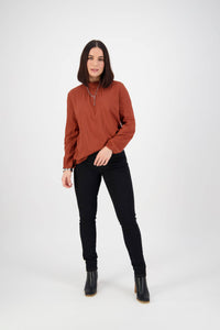 A woman with straight dark hair stands against a white background, wearing a rust-colored long-sleeve blouse, Vassalli Reign Jean in black denim, and black ankle boots. Her left hand is by her side as she looks at the camera.