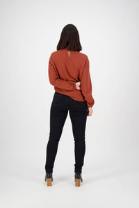 A woman faces away from the camera, wearing a rust long-sleeve top, Vassalli Reign Jean in Black Denim, and black heeled boots. She bends one arm behind her back against a plain white background.