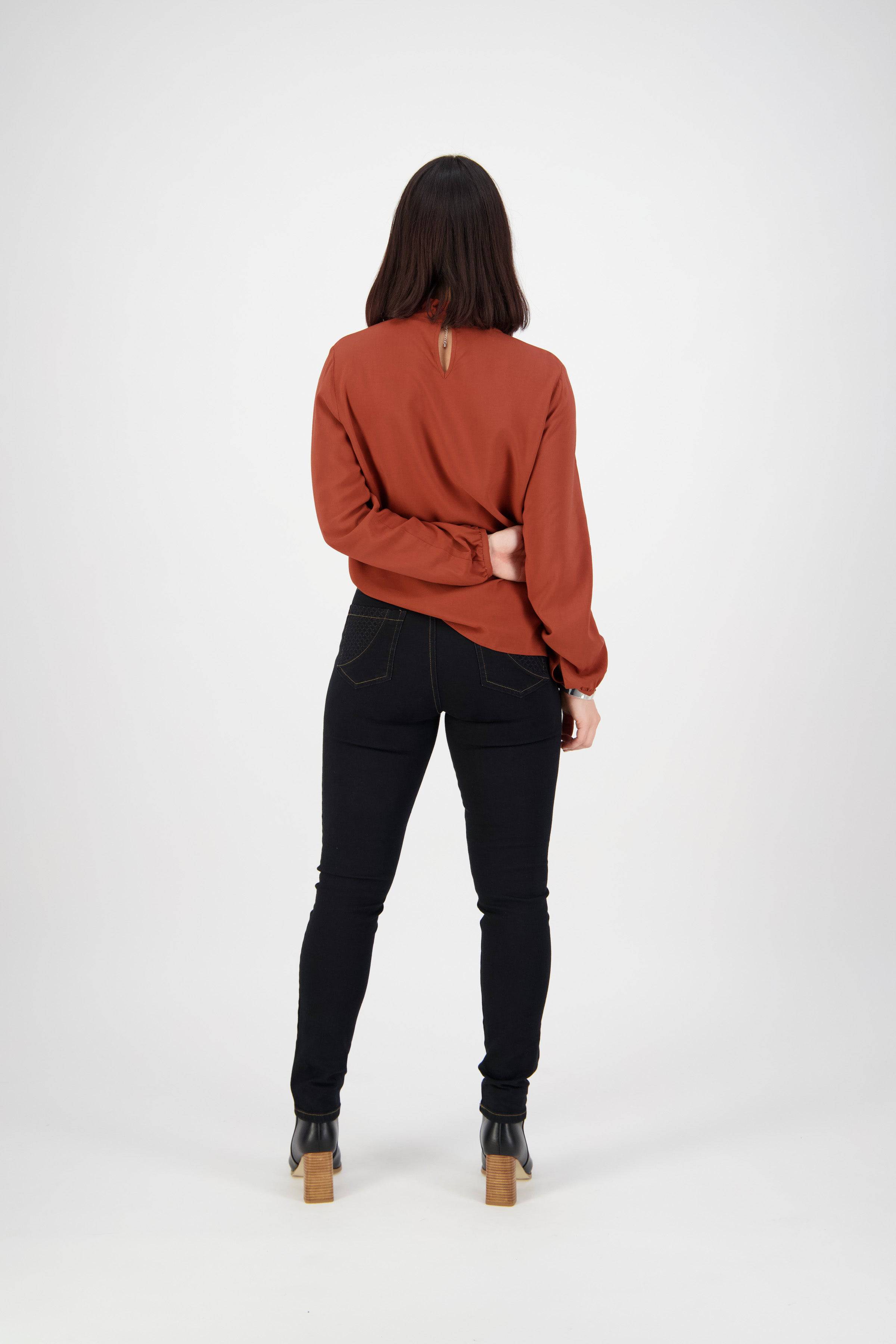 A woman faces away from the camera, wearing a rust long-sleeve top, Vassalli Reign Jean in Black Denim, and black heeled boots. She bends one arm behind her back against a plain white background.