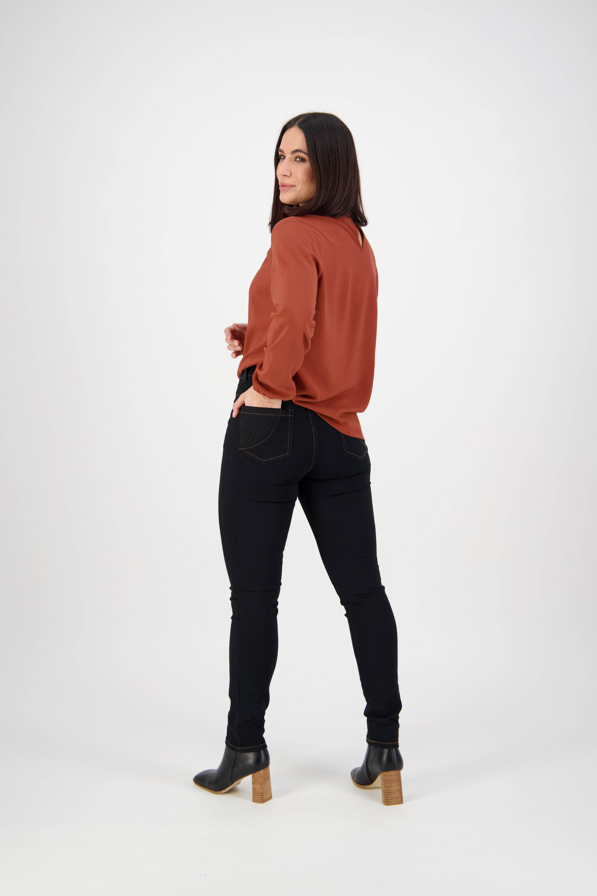 A woman with straight dark hair, wearing a rust long-sleeve blouse, Vassalli Reign Jean in black denim, and heeled black boots stands with one hand in her pocket and looks over her shoulder against a plain white background.