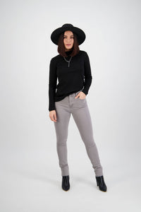 A woman wearing a black wide-brim hat, a black long-sleeve top, and Vassalli's Reign Jean in Light Grey stands against a plain white background with one hand in her pocket.
