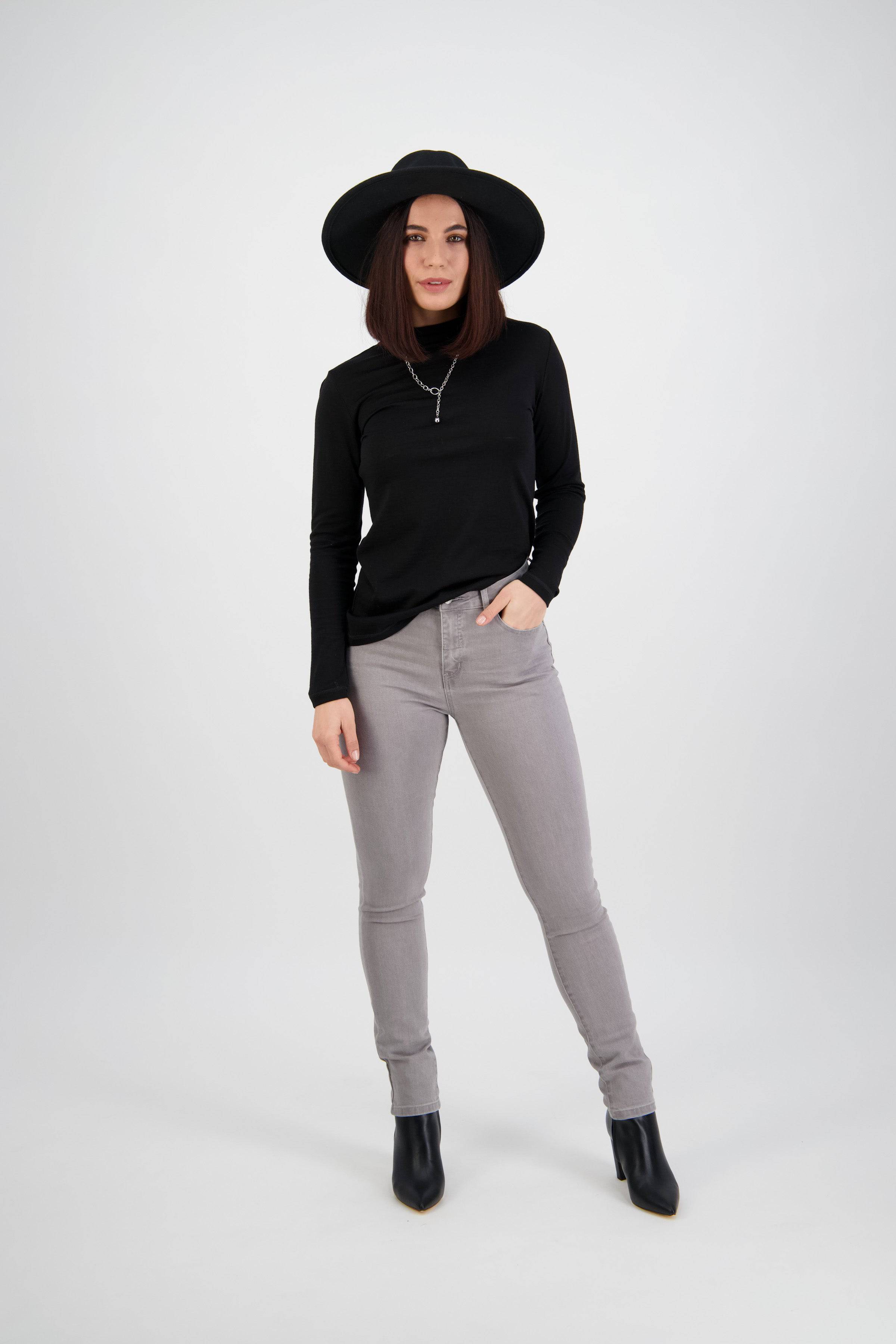 A woman wearing a black wide-brim hat, a black long-sleeve top, and Vassalli's Reign Jean in Light Grey stands against a plain white background with one hand in her pocket.