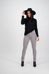 A woman stands in a studio against a plain white background, smiling and holding her black hat while wearing a black long-sleeve top, black boots, and Vassalli's Reign Jean in Light Grey.