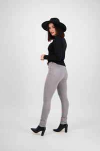 Woman in a black hat, black long-sleeve top, and Vassalli Reign Jean - Light Grey, paired with black heeled boots, stands against a plain light background looking over her shoulder.
