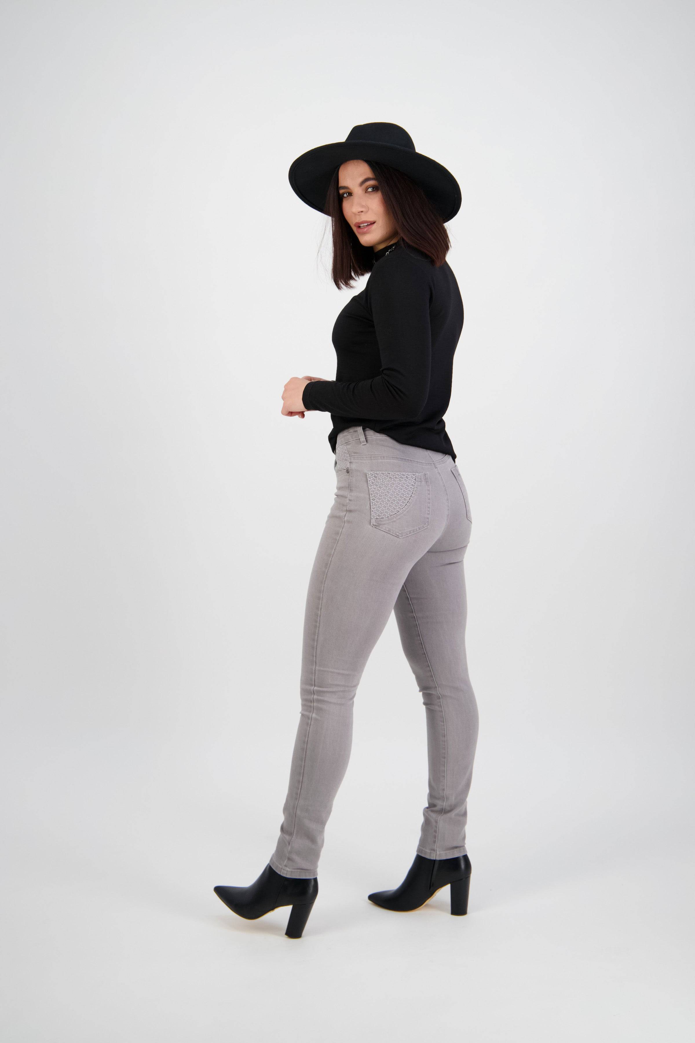 Woman in a black hat, black long-sleeve top, and Vassalli Reign Jean - Light Grey, paired with black heeled boots, stands against a plain light background looking over her shoulder.