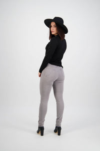 A woman stands facing away, looking over her shoulder. She wears a black wide-brim hat, black long-sleeve top, Vassalli Reign Jean in light grey, and black heeled boots against a plain white background.