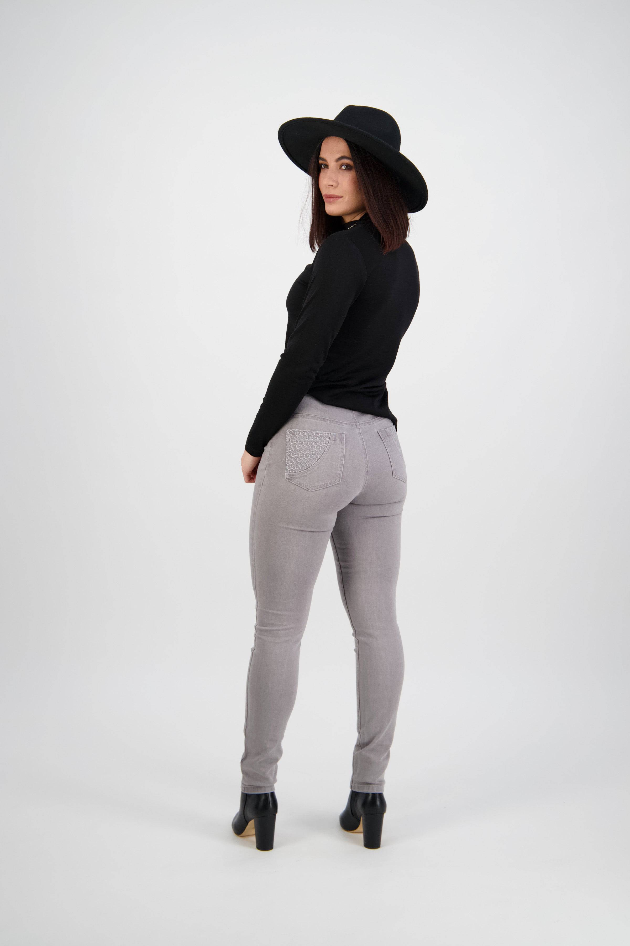 A woman stands facing away, looking over her shoulder. She wears a black wide-brim hat, black long-sleeve top, Vassalli Reign Jean in light grey, and black heeled boots against a plain white background.