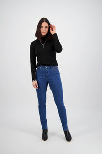 A woman with shoulder-length dark hair stands against a light background, wearing Vassalli's Reign Jean - Plain New Blue, a black long-sleeve top, black boots, and a necklace. She smiles softly and touches her hair with one hand.