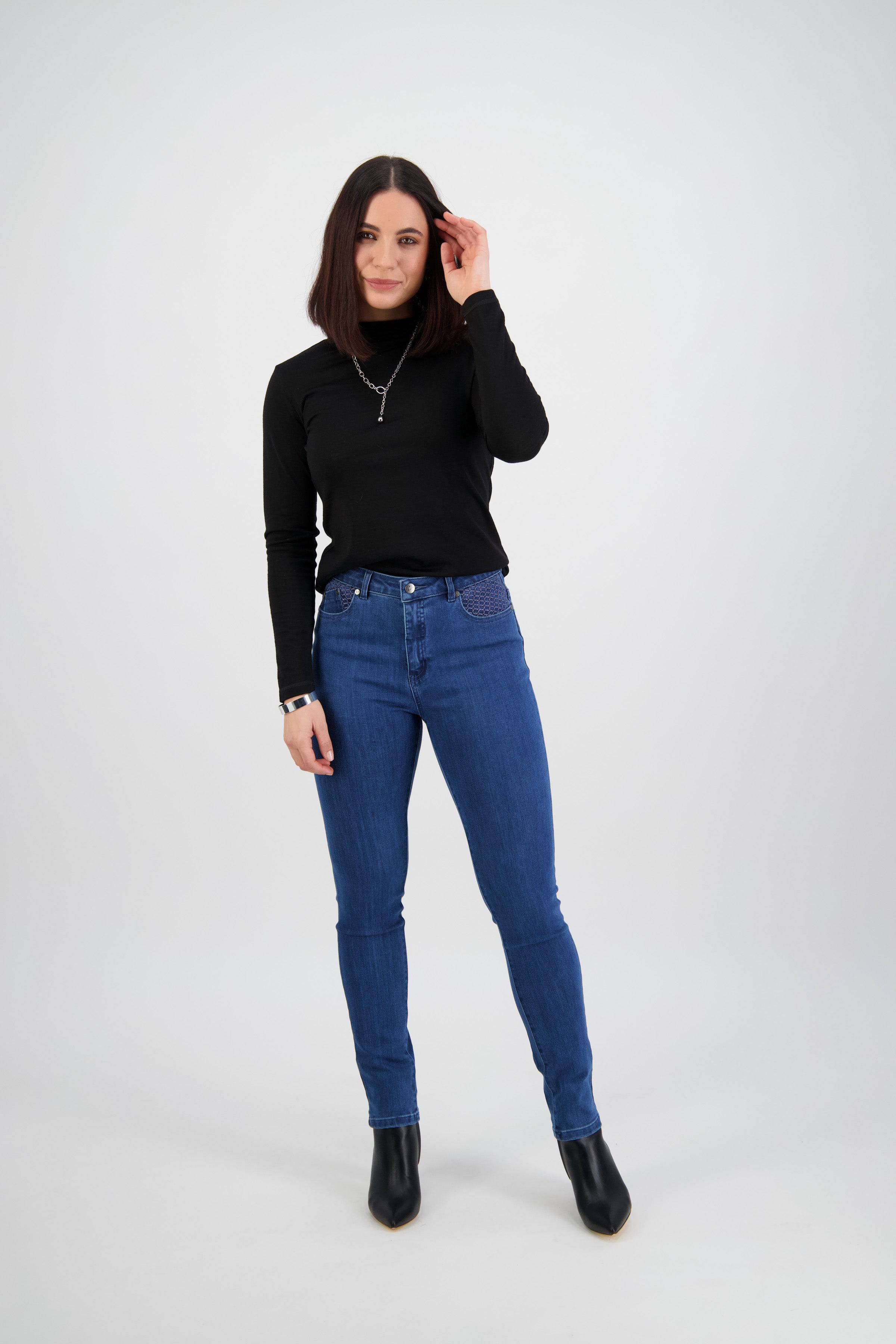 A woman with shoulder-length dark hair stands against a light background, wearing Vassalli's Reign Jean - Plain New Blue, a black long-sleeve top, black boots, and a necklace. She smiles softly and touches her hair with one hand.