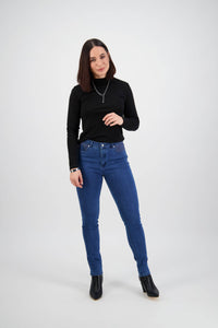 A woman with straight dark hair wears the Vassalli Reign Jean - Plain New Blue, a black long-sleeve top, black ankle boots, and a necklace. She stands against a plain white background, facing the camera with a slight smile.