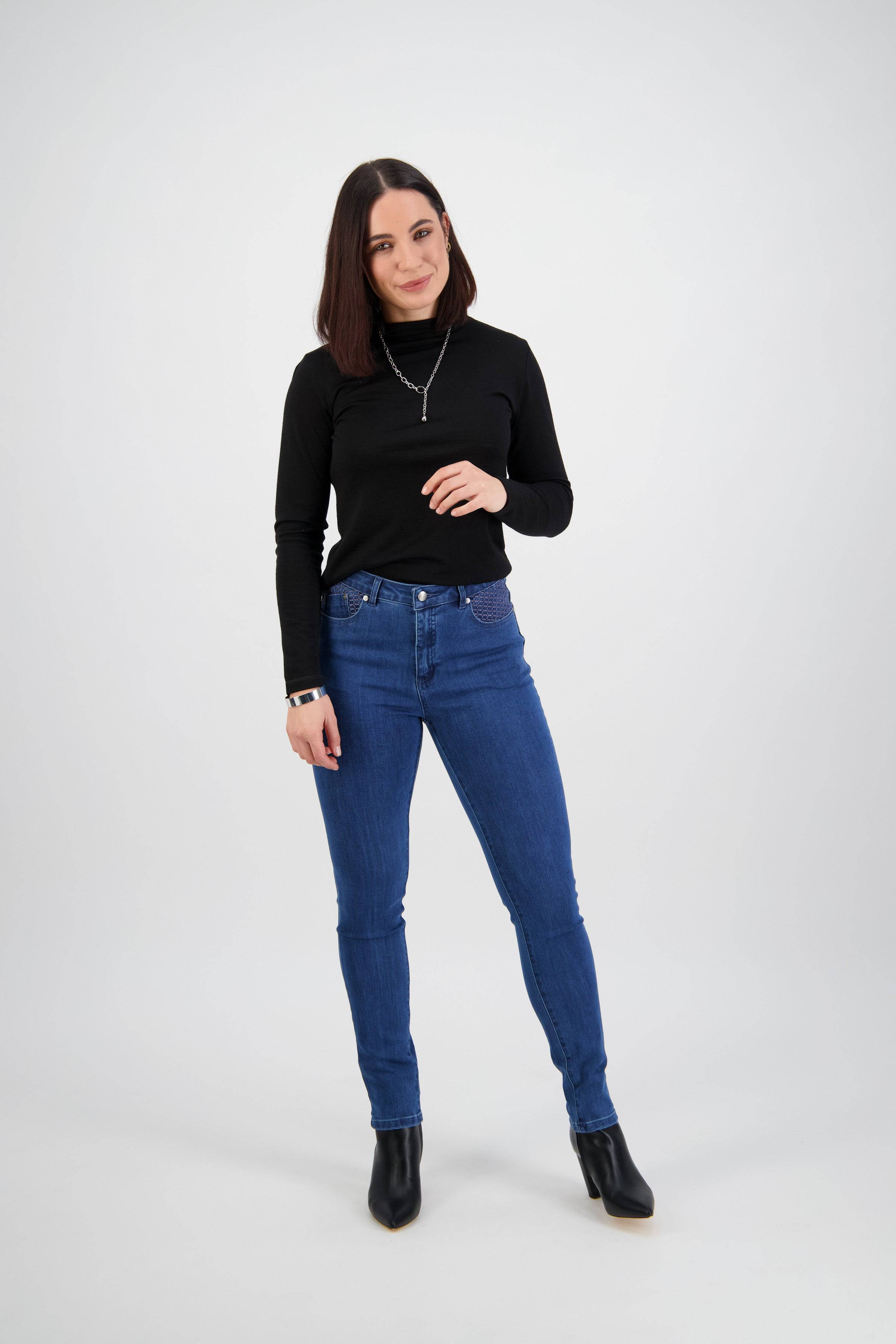 A woman with straight dark hair wears the Vassalli Reign Jean - Plain New Blue, a black long-sleeve top, black ankle boots, and a necklace. She stands against a plain white background, facing the camera with a slight smile.