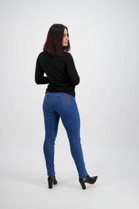 A woman with shoulder-length dark hair, wearing the Vassalli Reign Jean - Plain New Blue, a black long-sleeve top, and black heeled boots stands with her back to the camera, looking over her shoulder against a plain white background.