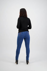 A woman with straight dark hair faces away, wearing a black top, Vassalli Reign Jean - Plain New Blue skinny jeans, and black heeled boots against a plain light gray background.