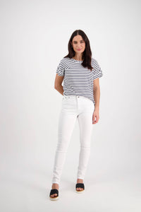 A woman with long dark hair wears a black-and-white striped shirt, Vassalli Reign Jean - White Denim, and black sandals. She stands in a studio with a plain white background, smiling gently with one hand behind her back.