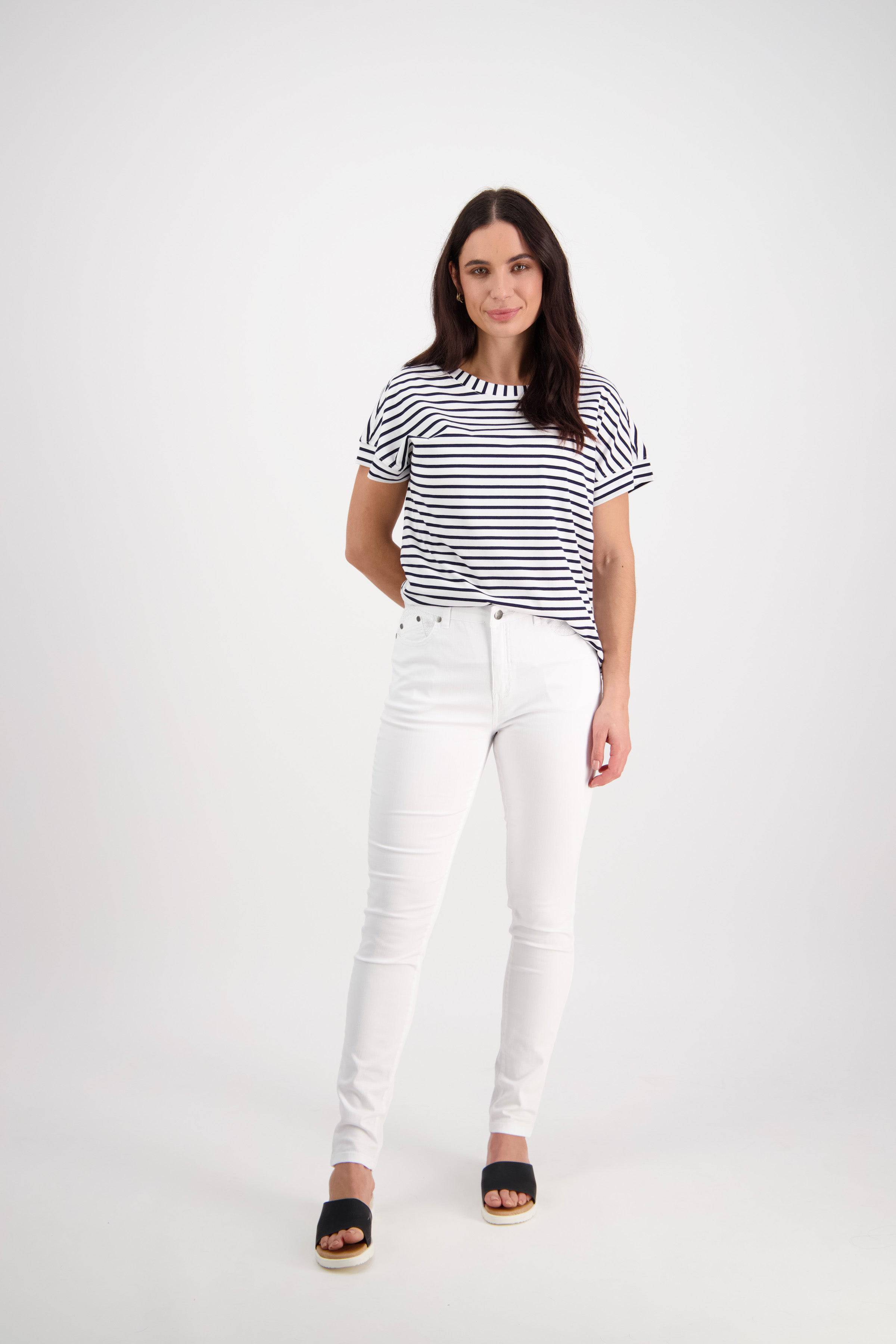 A woman with long dark hair wears a black-and-white striped shirt, Vassalli Reign Jean - White Denim, and black sandals. She stands in a studio with a plain white background, smiling gently with one hand behind her back.