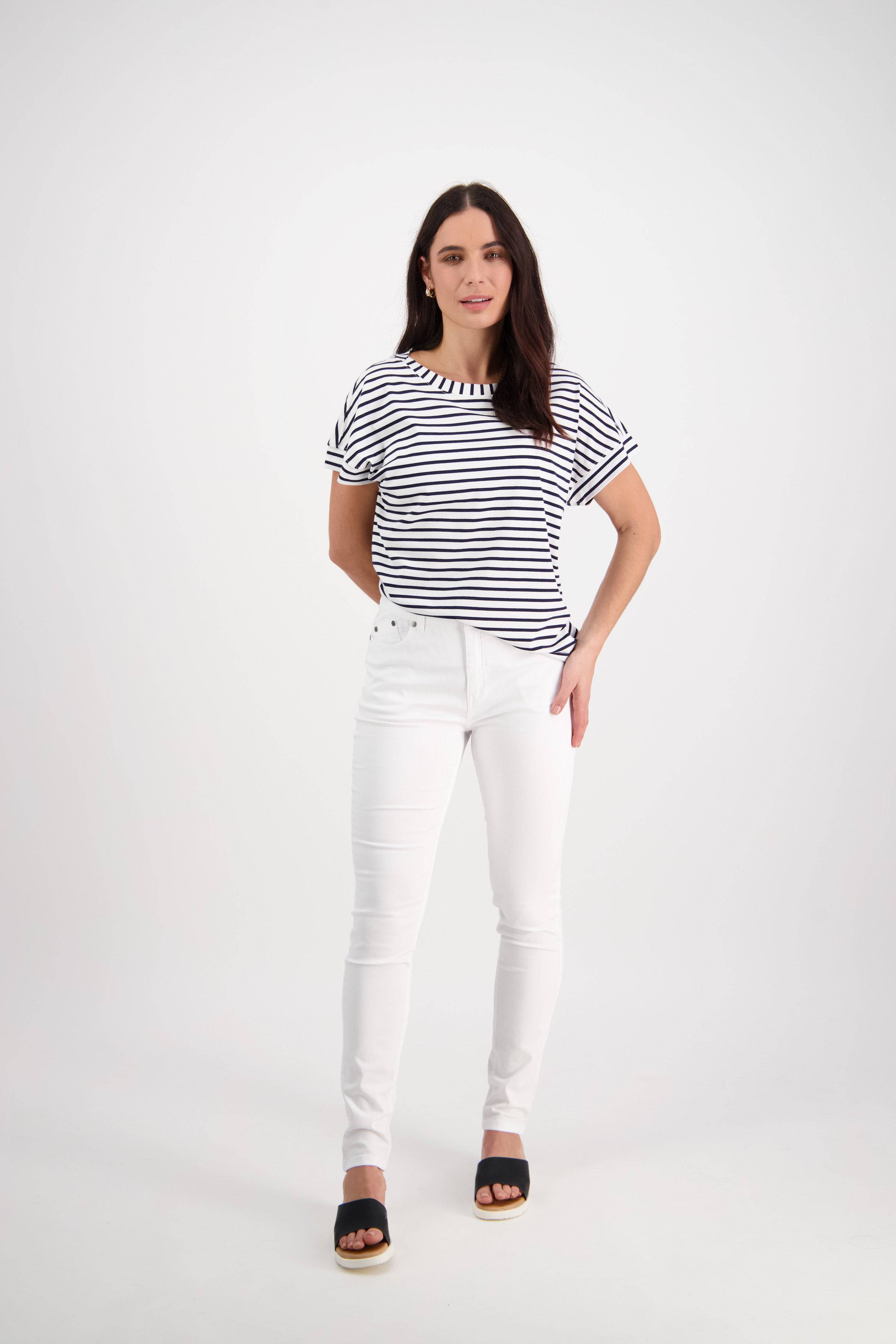A woman with long dark hair stands against a white background, wearing a black and white striped short-sleeve top, Vassalli Reign Jean in white denim, and black open-toe sandals. She faces forward with a slight smile.