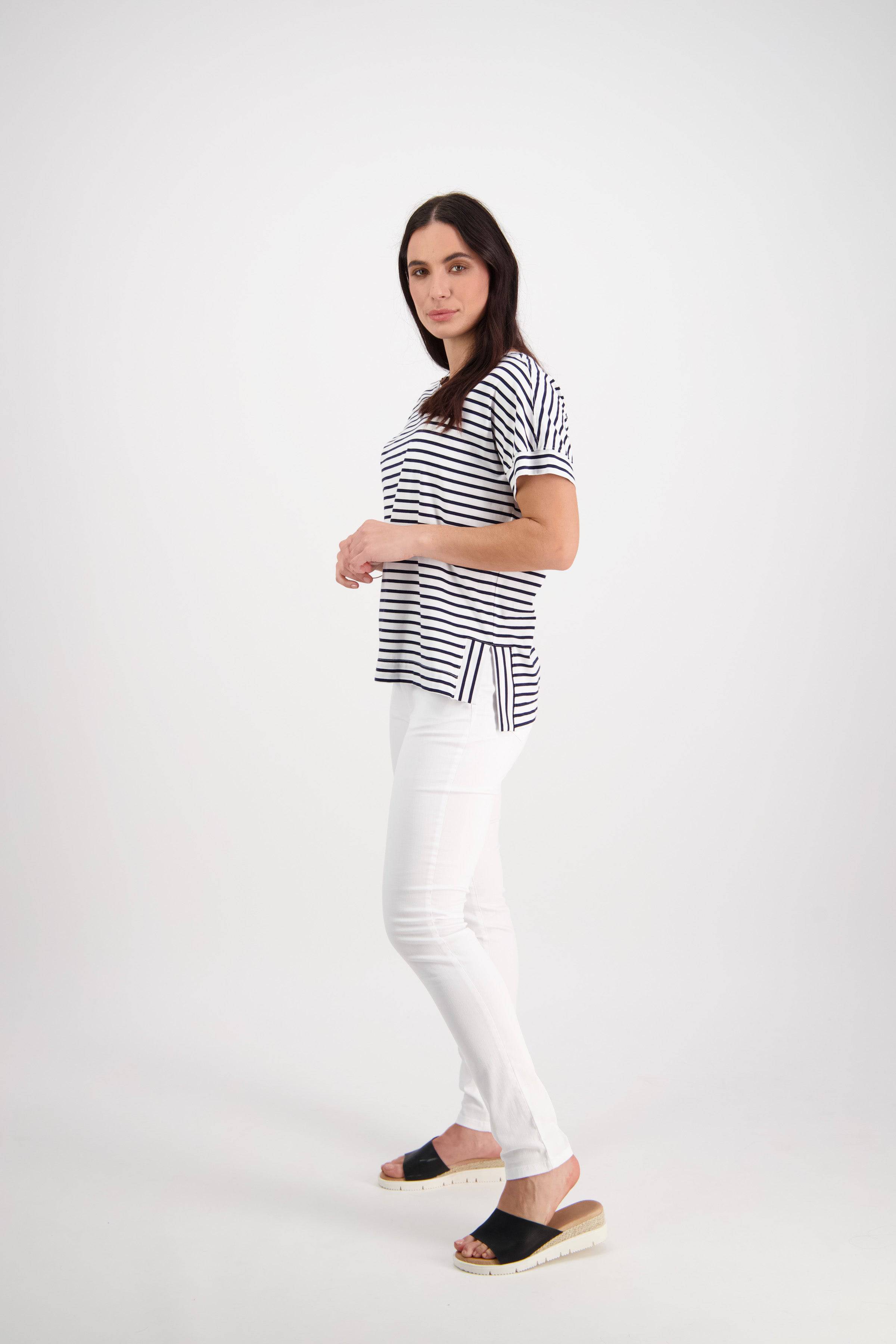 A woman with long dark hair stands in a studio, wearing a black-and-white striped shirt, Vassalli’s Reign Jean in white denim, and black slide sandals. She is facing slightly sideways against a plain white background.