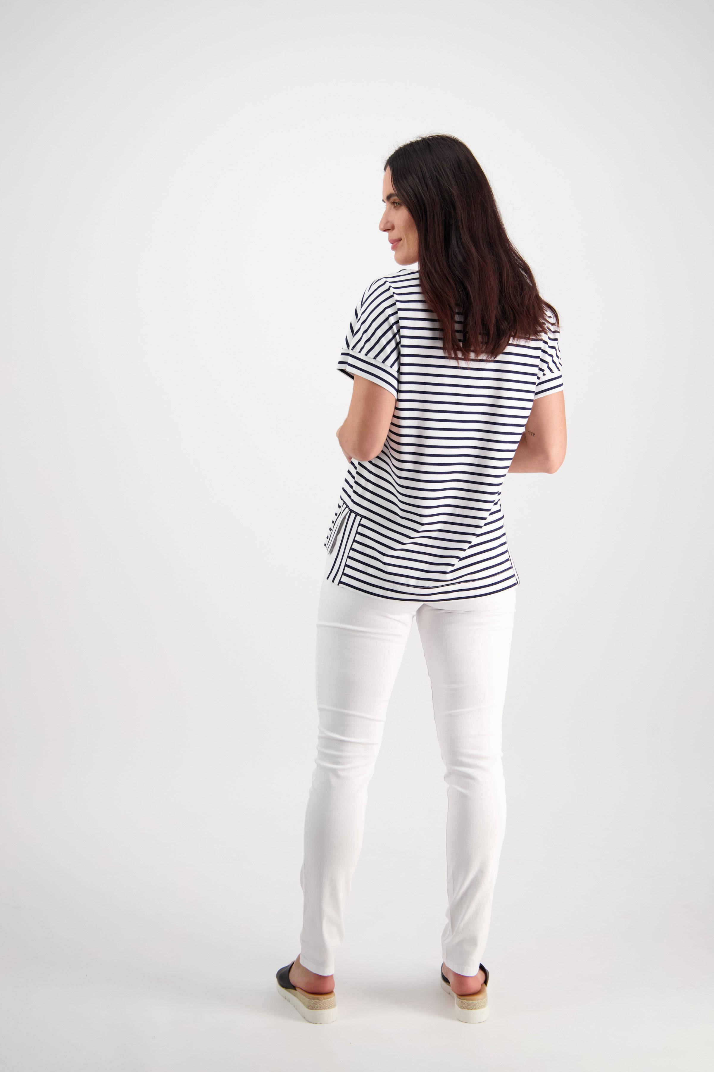 A woman with long dark hair stands facing slightly away, wearing a black and white striped short-sleeve top, Vassalli Reign Jean in White Denim, and slip-on shoes against a plain white background.