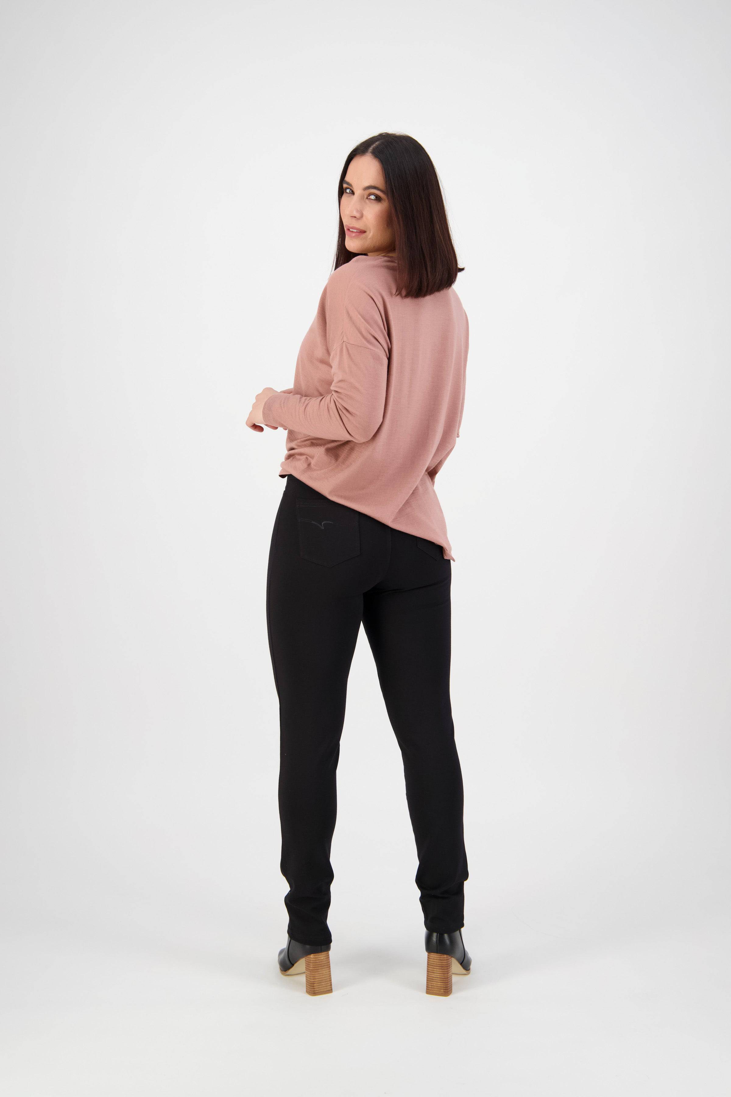 A woman with shoulder-length dark hair faces backward, head turned to the camera. She wears a pink long-sleeve top and Vassalli Ponti Pant - Black, set against a plain white background.