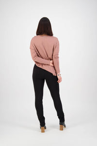 A woman faces away from the camera against a white background, wearing a light brown long-sleeve top, black heeled boots, and Vassalli's Ponti Pant - Black. One arm is bent behind her back, holding her other arm.