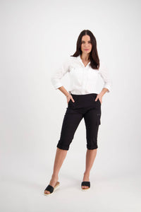 A woman stands confidently against a plain white background wearing the Vassalli Wave Pant in black and a white button-up shirt, paired with black slip-on sandals. She has long dark hair and her hands rest on her hips.