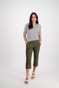 A woman with long dark hair stands against a white background wearing the Vassalli Wave Pant in Khaki with an adjustable waistband, paired with a black-and-white striped top and white sandals. Her hands are in her pockets.