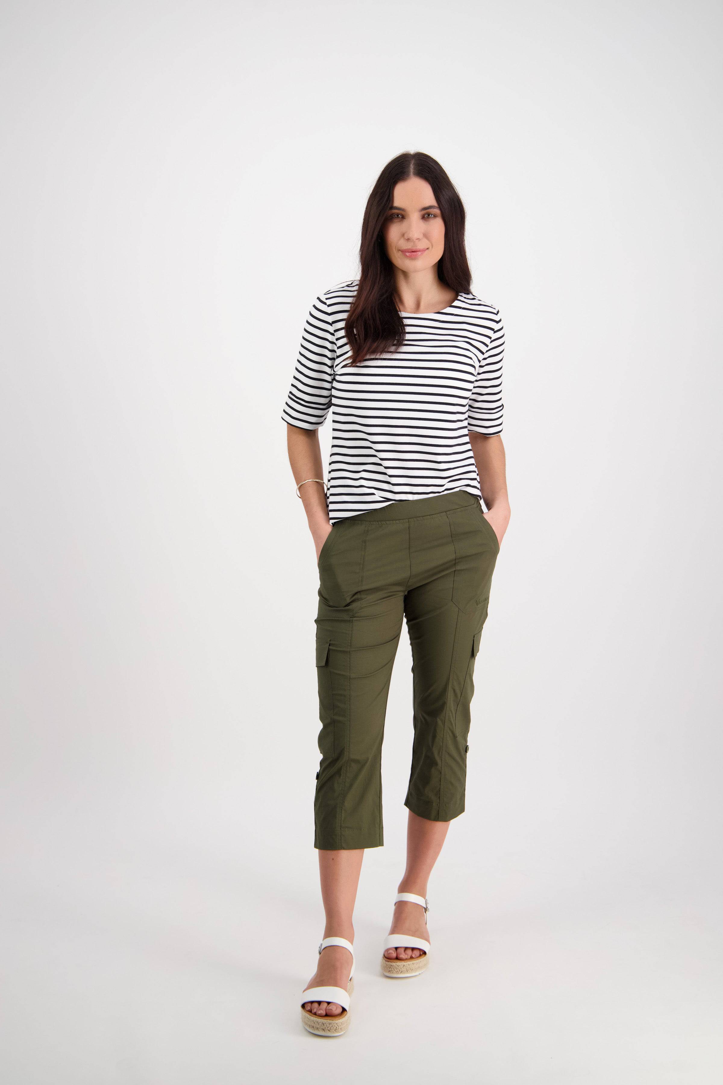 A woman with long dark hair stands against a white background wearing the Vassalli Wave Pant in Khaki with an adjustable waistband, paired with a black-and-white striped top and white sandals. Her hands are in her pockets.