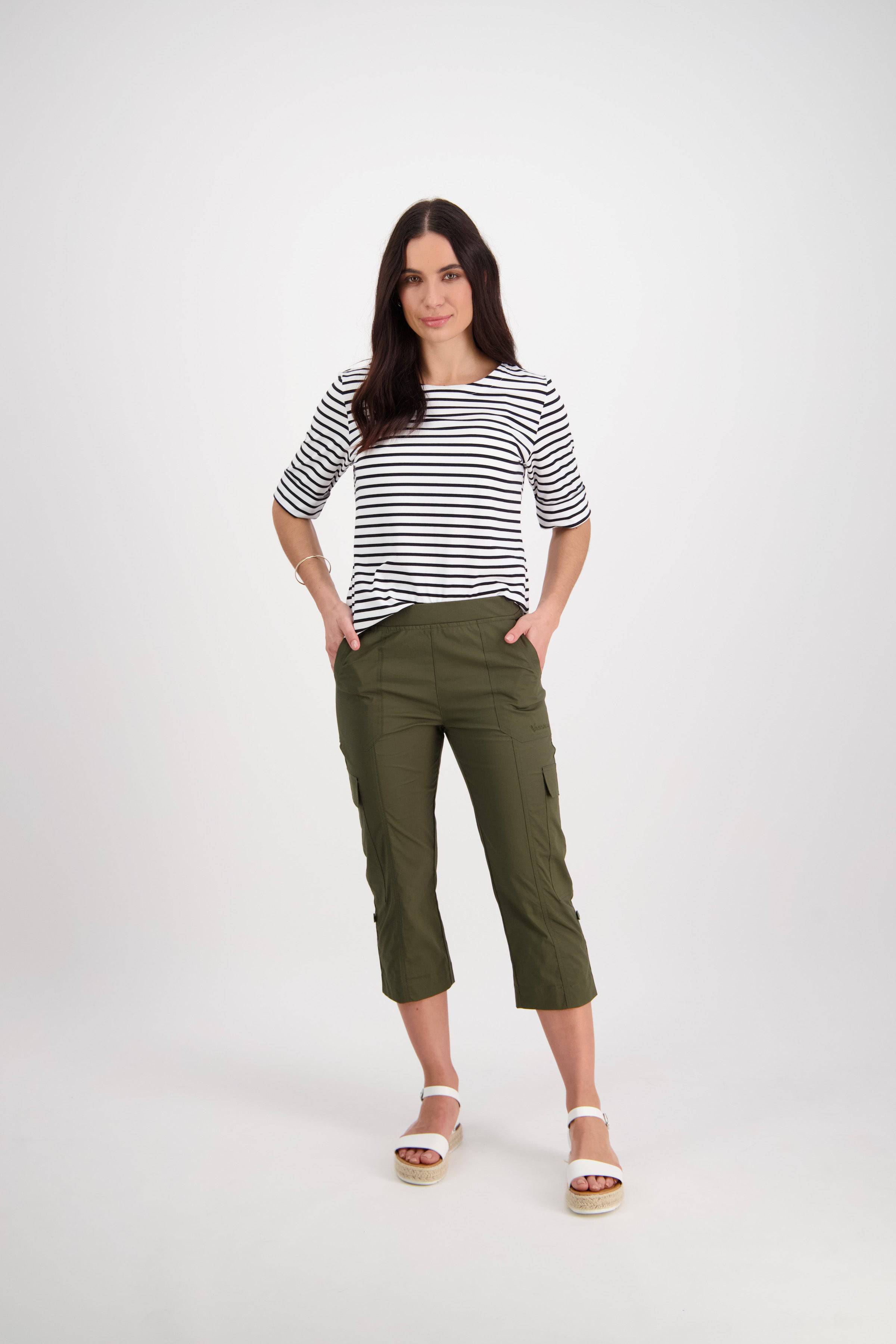 A woman with long dark hair stands confidently against a white background wearing Vassalli's Wave Pant - Khaki, relaxed cargo pants in stretch poplin with an adjustable waistband. She pairs them with a black-and-white striped top and has her hands in her pockets.