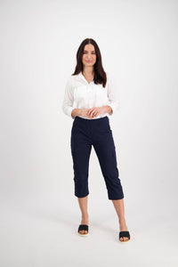 A woman with long dark hair smiles against a white background, wearing a white shirt, Vassalli Wave Pant - Navy with pockets and adjustable waistband, and black slide sandals.